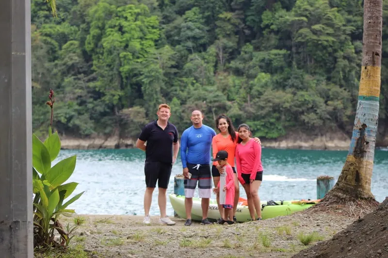 Five people stand beside a kayak on the shore of a cove ringed by forested hills.
