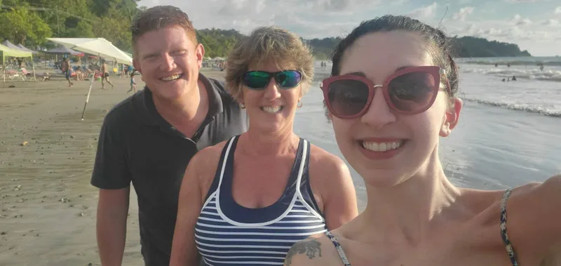 Three people lean in for a selfie on the beach, with a forested point behind them.