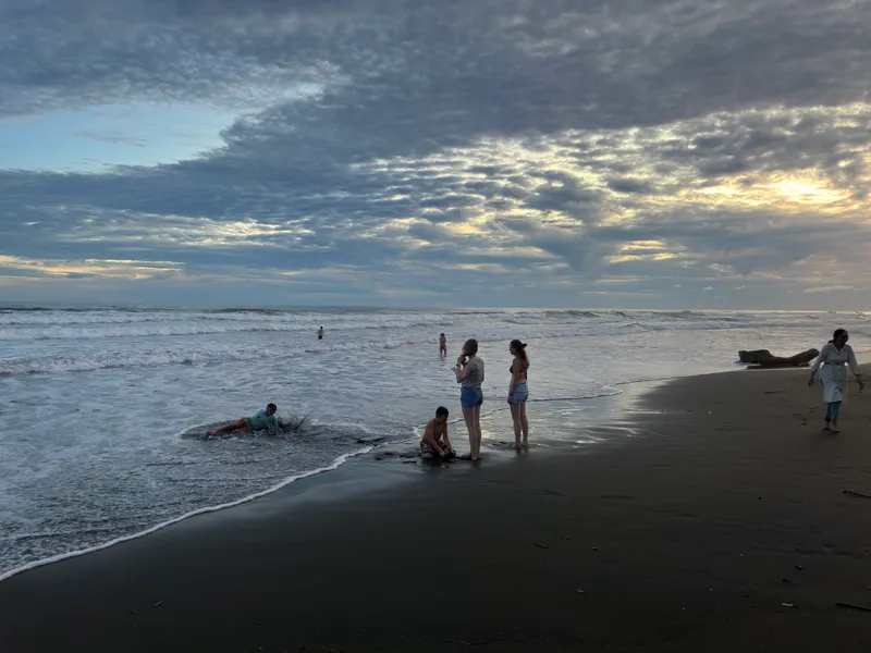 People standing and swimming in the shallow surf at dusk, under a wide cloudy sky.