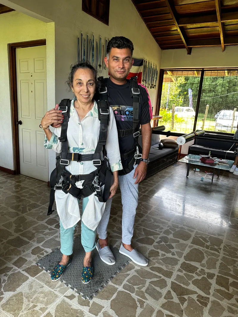 A woman and a man wearing skydiving harnesses stand together in a tiled room.