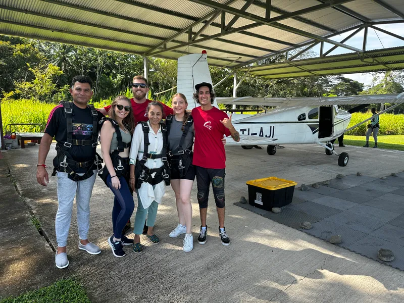 Six people in harnesses pose beside a small white plane under a metal roof.