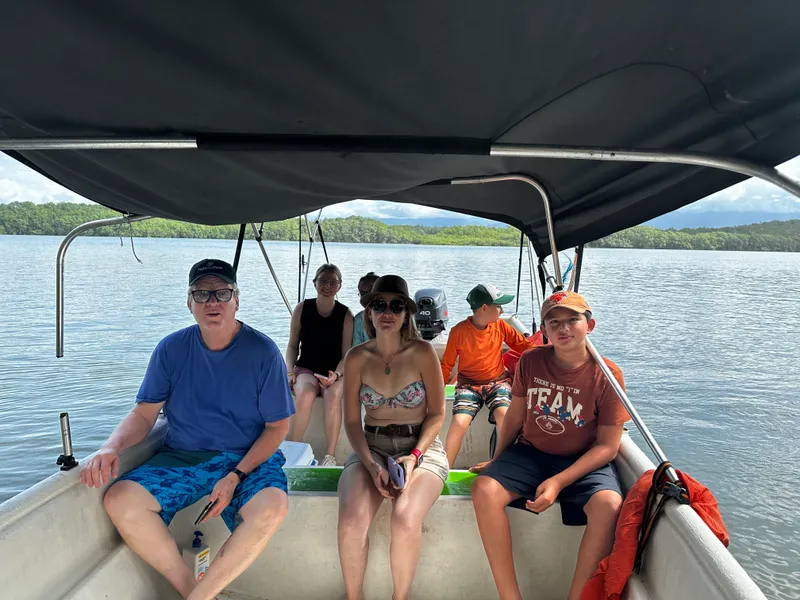 Five people sit under the canopy of a boat on calm water, with forest along the far shore.