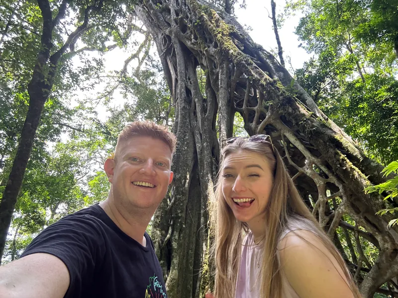 Two people take a selfie in front of a big tree with roots hanging down around its trunk.