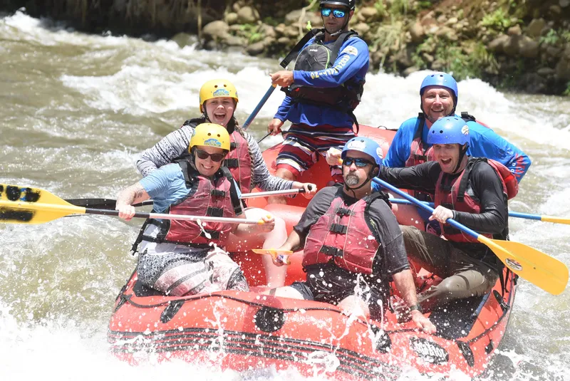 A crew in helmets and life jackets paddles a red raft through a rapid, water breaking over the bow.