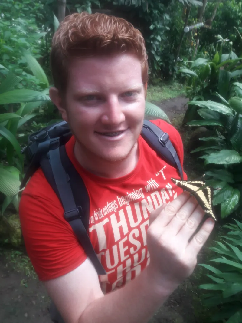 A man in a red shirt holds a butterfly resting on his open hand, green leaves behind him.