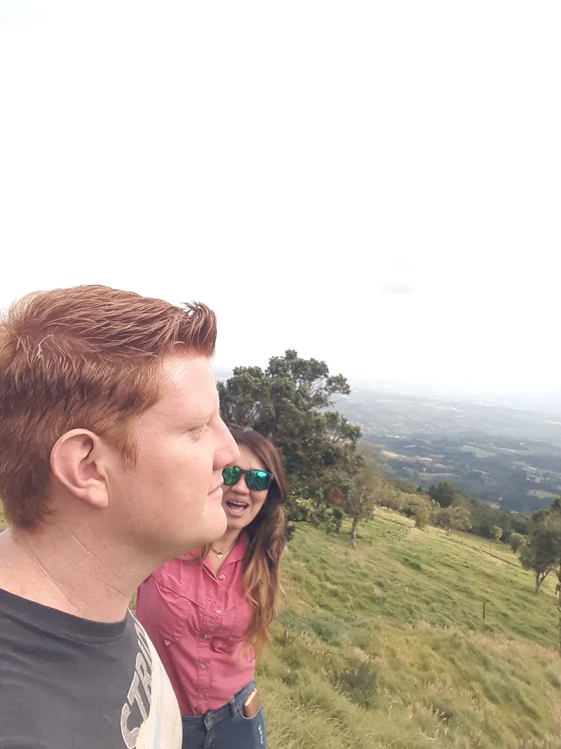 Two people stand on a ridge with pasture, scattered trees and a hazy valley below.