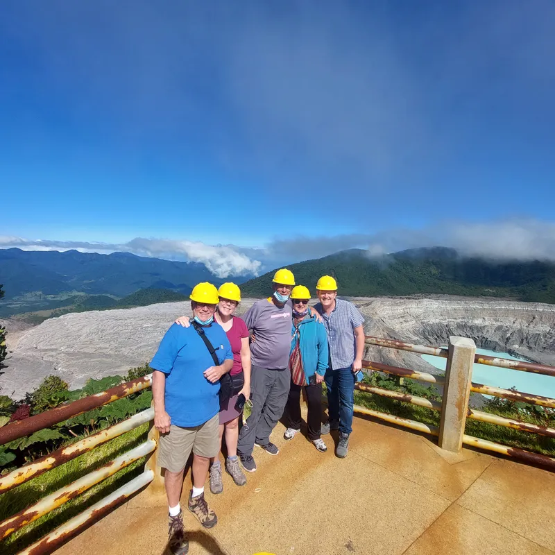 Six visitors in yellow hard hats stand on a lookout above a wide crater with a pale blue lake.