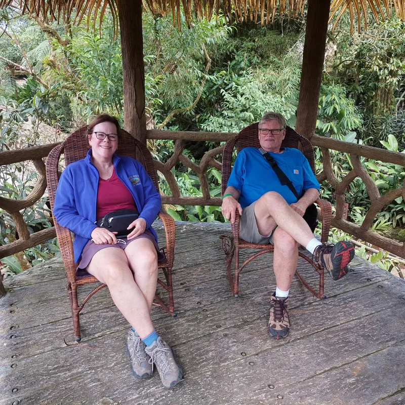 Two visitors sit in wicker chairs on a wooden deck under a thatched shelter, dense greenery behind.