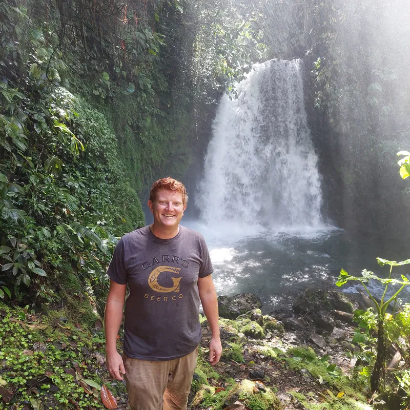 A man in a grey t-shirt stands on wet ground in front of a waterfall falling over a dark cliff.