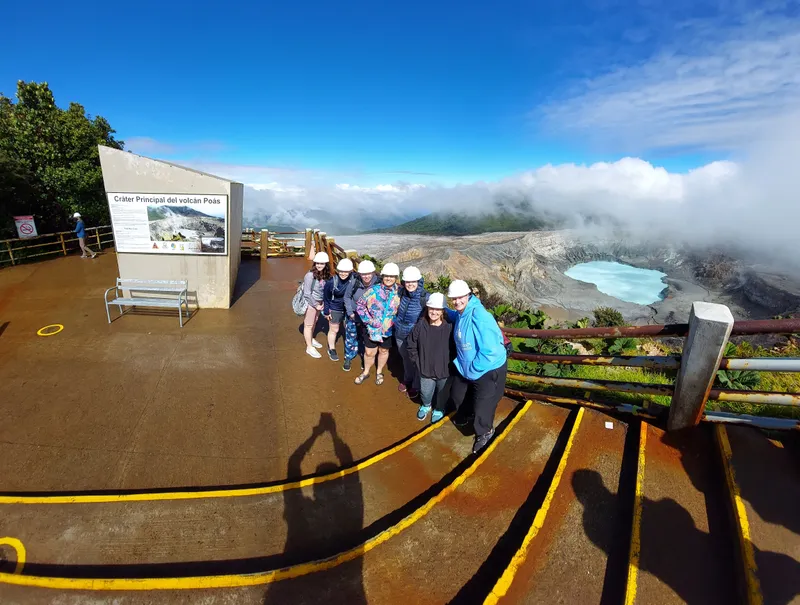 A line of visitors in white hard hats poses at a railing next to an information sign, with a crater and its lake behind.