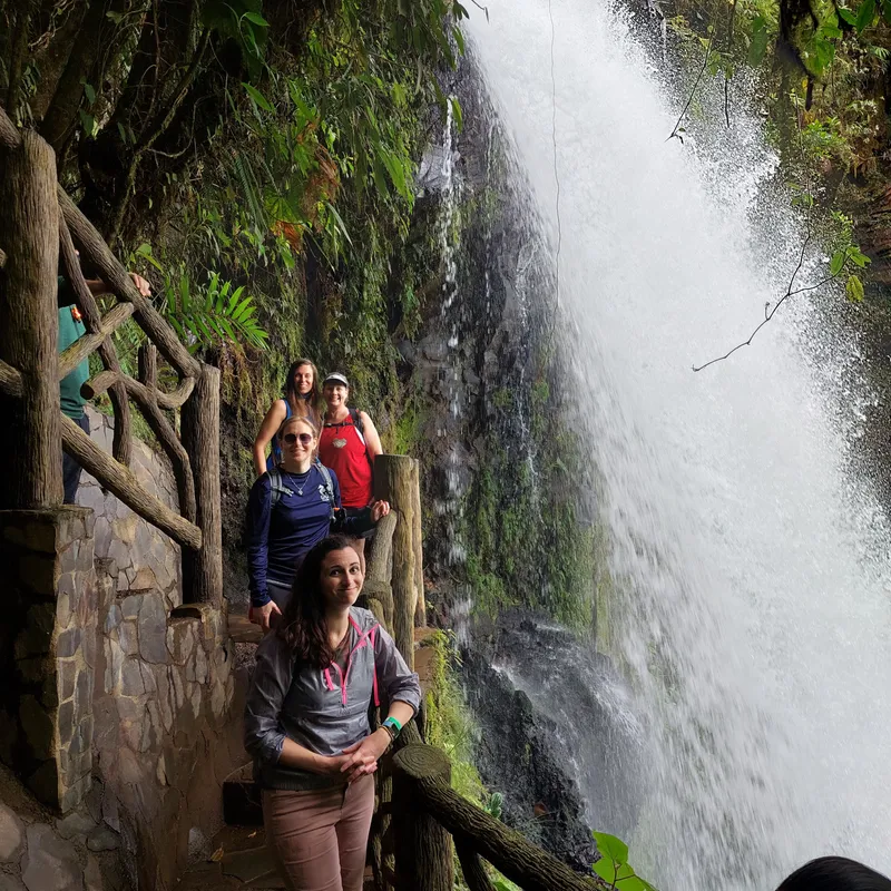 Three people stand on a wooden stairway beside a tall waterfall running down a mossy wall.