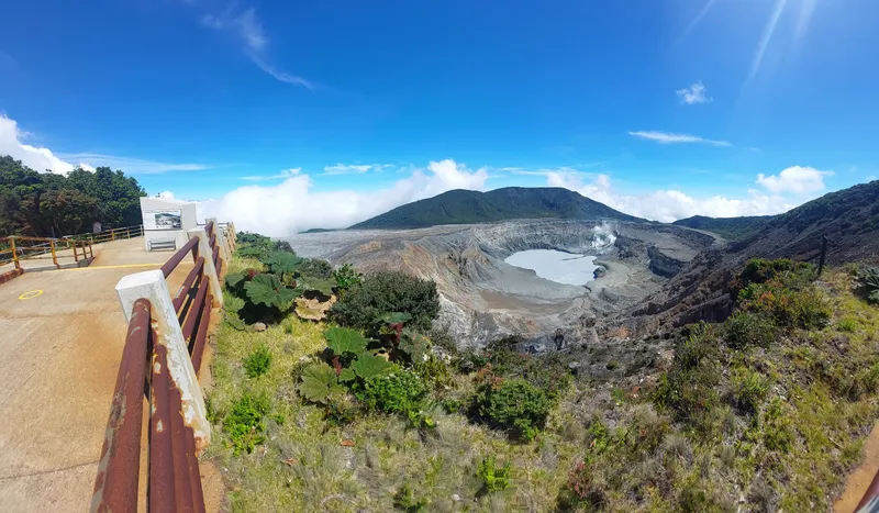 A crater holding a green blue lake, seen from a fenced viewpoint on the rim.