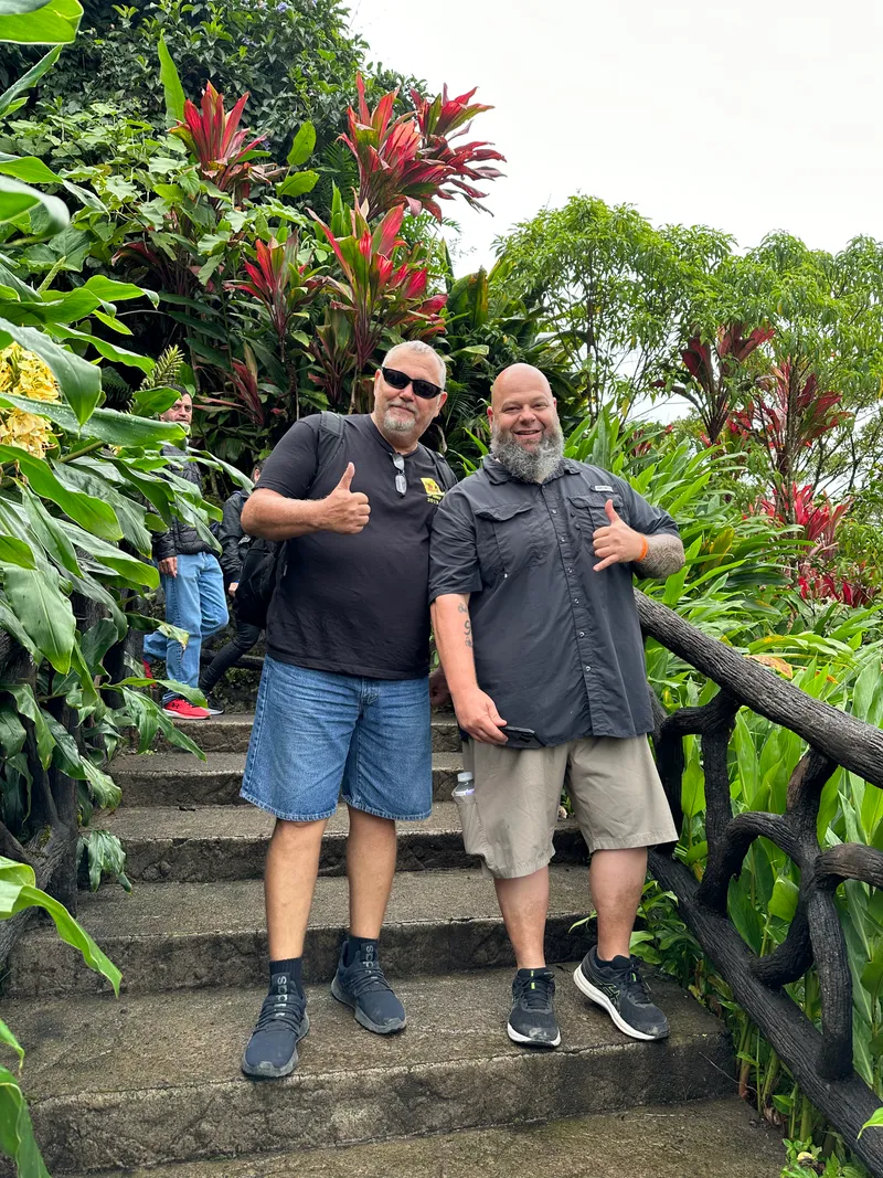 Two men give a thumbs up on stone steps surrounded by red and green tropical plants.