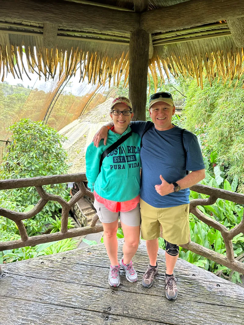 A couple poses under a thatched roof at a railing, with a waterfall on the slope behind.