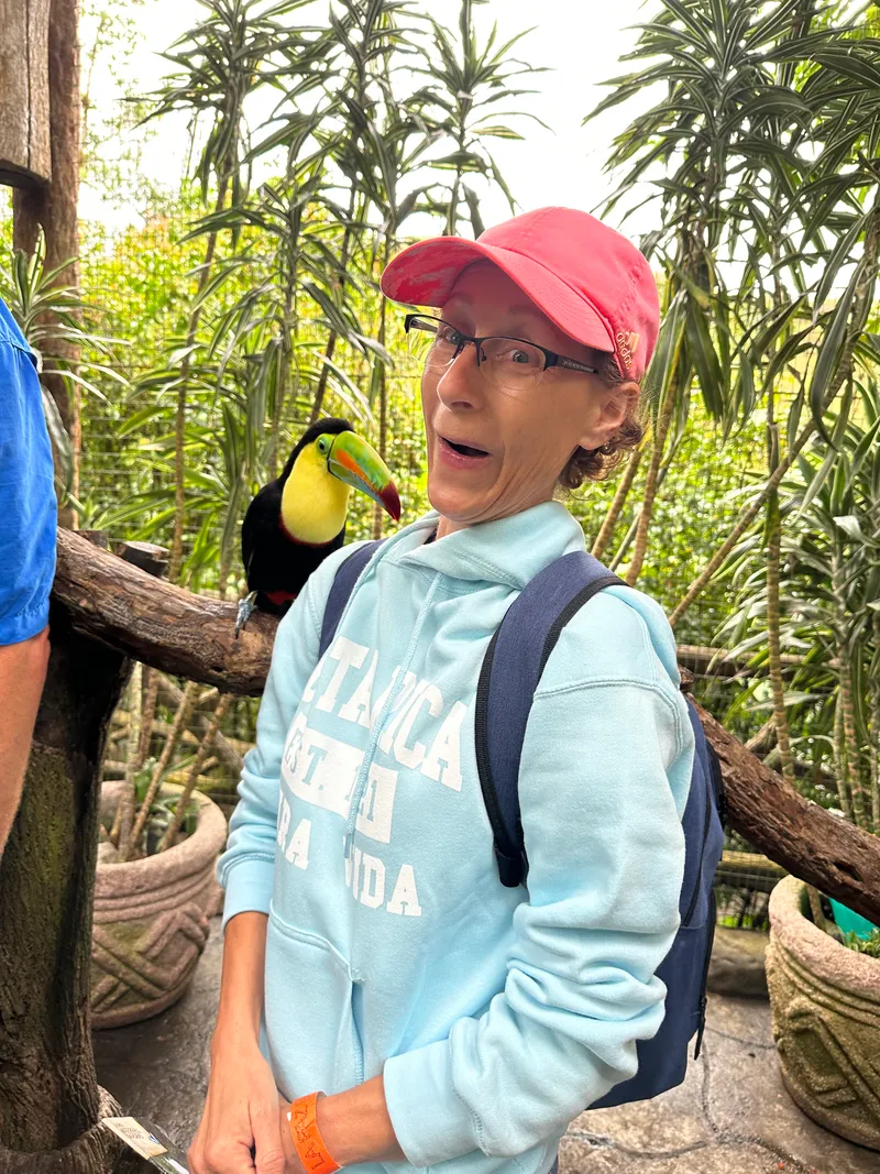 A woman in a red cap smiles beside a toucan perched on a branch.