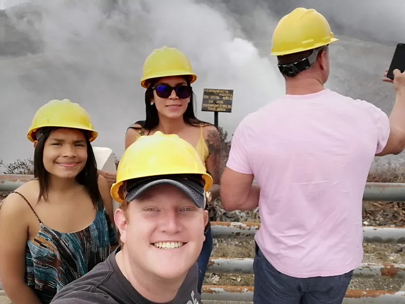 Four visitors in yellow hard hats near a sign, steam rising from the ground behind them.