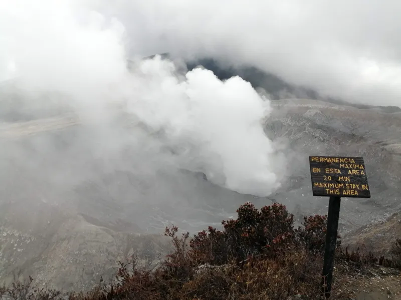 A thick column of white steam rises from grey crater ground next to a warning sign.