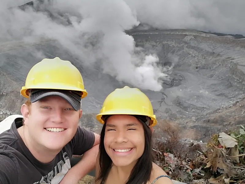 Two people in yellow hard hats take a selfie with steam and the crater wall behind them.