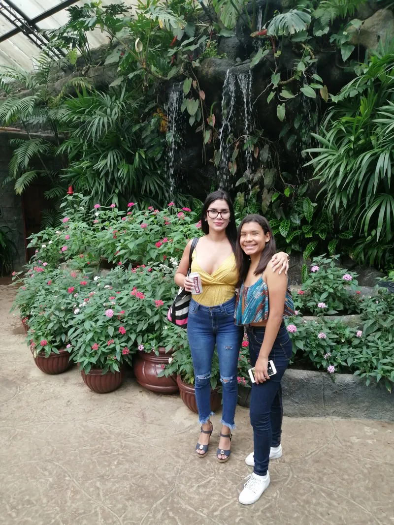 Two women stand among potted flowering plants with a waterfall behind them.