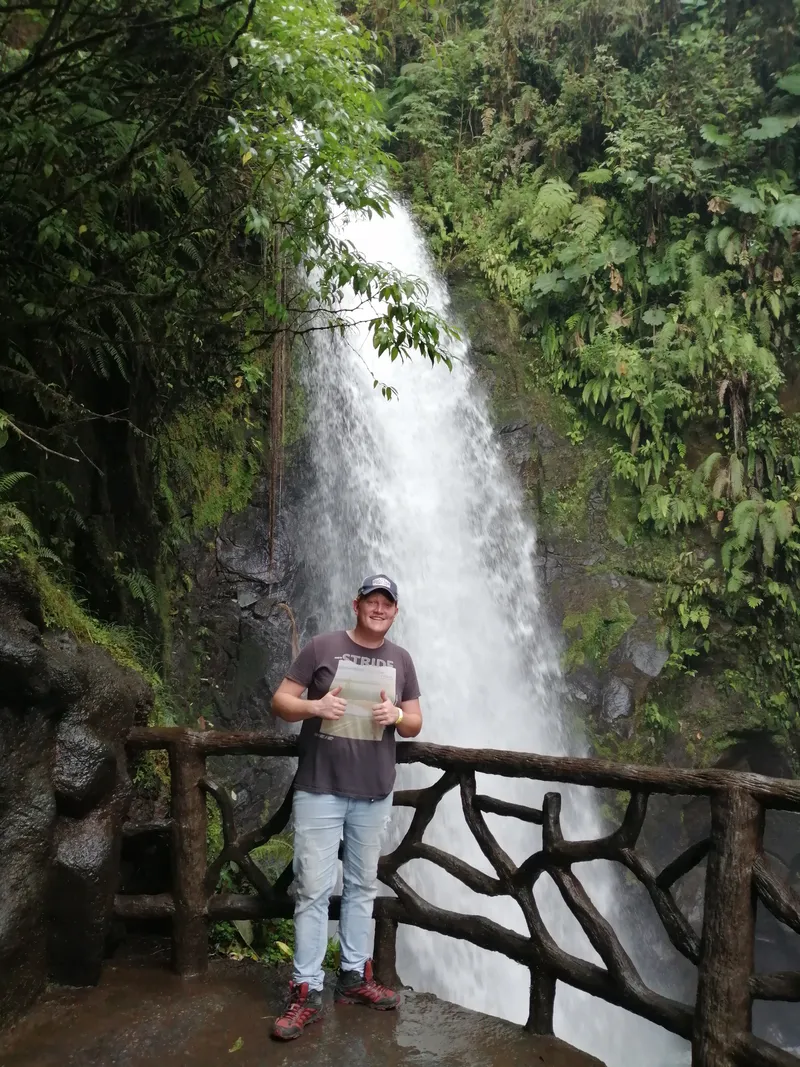 A man stands on a wooden viewing deck in front of a tall waterfall.