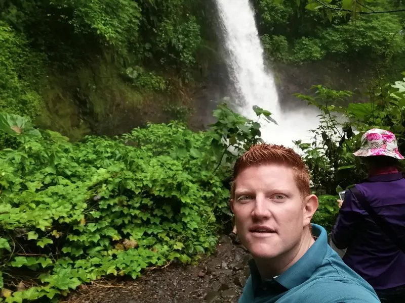 A man takes a selfie with a waterfall and dense green foliage behind him.