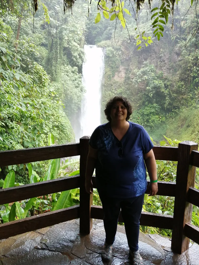 A woman stands at a wooden railing with a tall waterfall behind her.