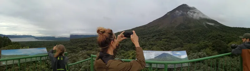 Someone raises a phone to photograph the slope of a volcano from a lookout.