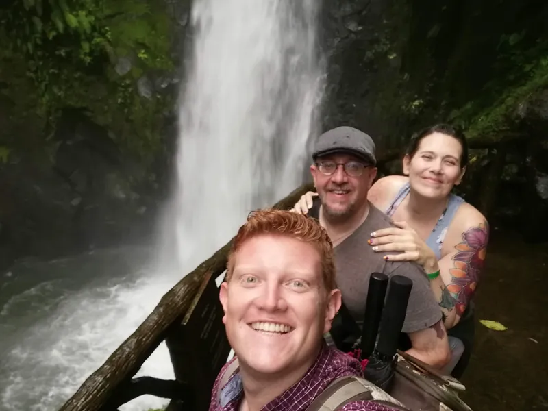 Three people crowd into a photo at a wooden railing in front of a waterfall.