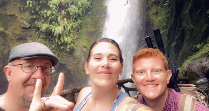 Two people take a selfie in front of a waterfall, one making a peace sign.