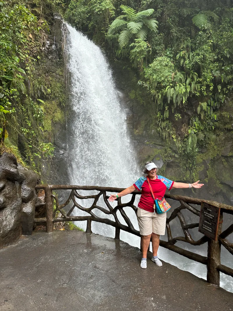A woman opens her arms at a railing in front of a waterfall.