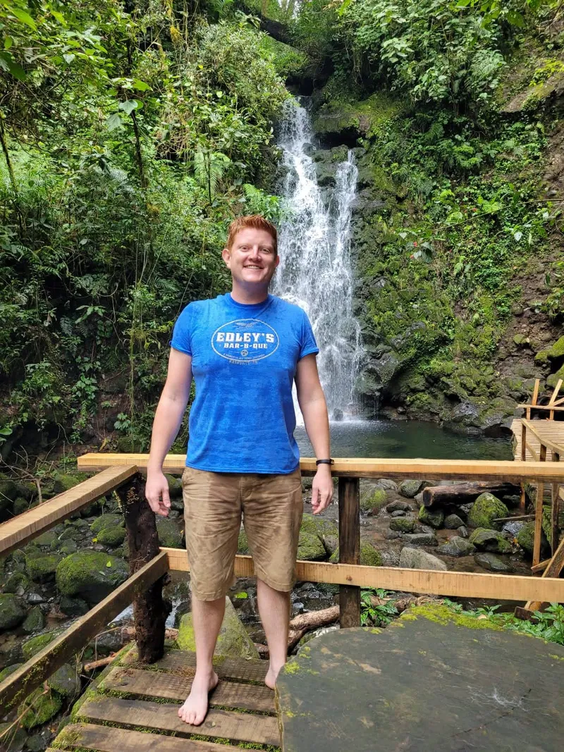 A man in a blue t-shirt stands on a wooden walkway in front of a waterfall in the forest.